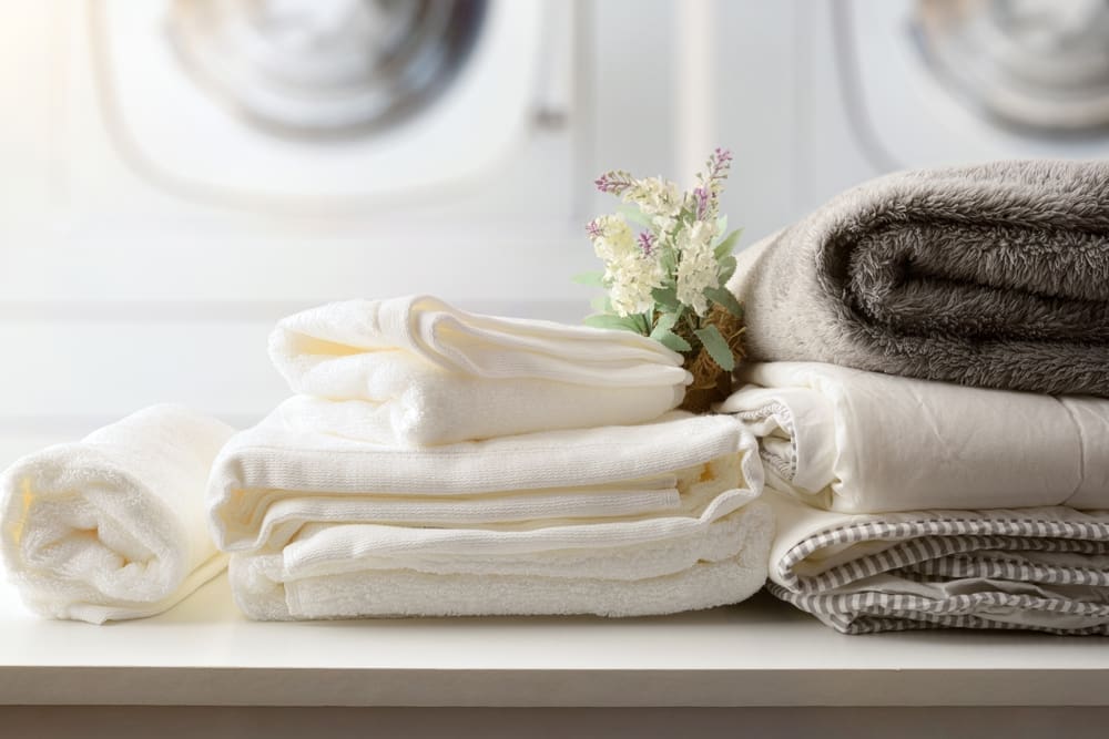 Stacked clean towels and linens, including white and beige fabrics, are neatly folded on a countertop with a small decorative bouquet, set in a laundry room.