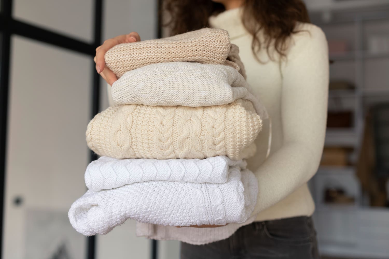 A person holding a stack of neatly folded cream and white knit sweaters, standing indoors, shows the fresh results of a wash and fold laundry routine.