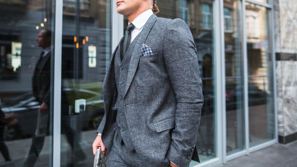 A man in a grey suit, white shirt, and striped tie is walking outside a modern glass building, holding a folder or notebook.