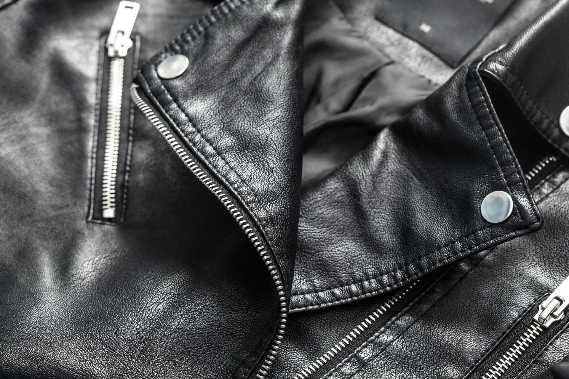 Close-up of a black leather jacket showing silver zippers, snap buttons, and textured material with visible stitching.